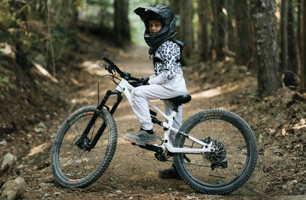 Child on a full-suspension kids mountain bike on a forest trail, wearing a helmet and MTB gear